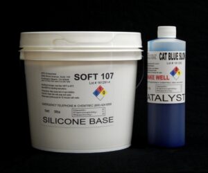 A white plastic bucket labeled "10 lb Kit : Soft 107 – (7 shore A durometer) – Longer Pot Life Mold Making Silicone" and a blue plastic bottle labeled "cat blue slg catalyst" against a black background.