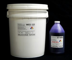 A white plastic bucket labeled "44 lb Kit : Medium 122 – (22 shore A durometer) – Regular Pot Life Mold Making Silicone" next to a purple bottle of catalyst marked "shake well." both items are displayed against a black background.