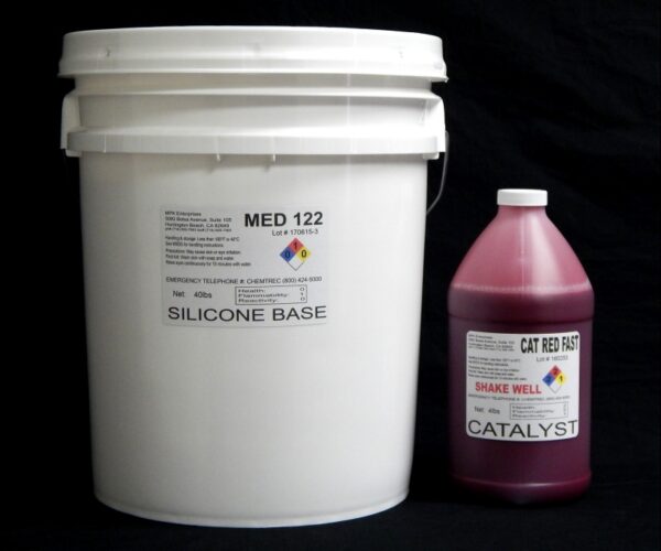 A white industrial bucket labeled "44 lb Kit : Medium 122 – (22 shore A durometer) – Fast Curing Mold Making Silicone" next to a pink bottle labeled "cat red fast catalyst" against a black background.