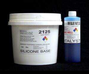 Two chemical containers: a white bucket labeled "10 lb Kit :#2125 Base w/ MM Blue Fast Catalyst – (25 shore A durometer)" and a blue bottle labeled "catalyst," both with hazard symbols and text details on a black background.