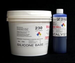 Two containers of chemicals: a large white tub labeled "11 lb Kit : QM 230 Blue Cat: 30 shore A – Platinum Silicone base" and a smaller blue bottle labeled "catalyst," both displaying hazard symbols, against a black background.