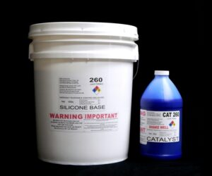 A white bucket labeled "Food Grade QM 260 FG B : 60 shore A – Platinum Silicone" and a blue bottle labeled "Blue Catalyst" on a black background, both featuring warning labels.