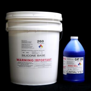 A white bucket labeled "Food Grade QM 260 FG B : 60 shore A – Platinum Silicone" and a blue bottle labeled "Blue Catalyst" on a black background, both featuring warning labels.