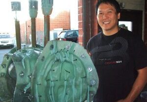 A man smiling at the camera, standing next to a large green mechanical part in a parking lot.